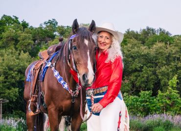 Smiling woman in western attire with a saddled horse in a green field.