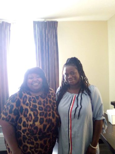 Two women smiling indoors beside a table.