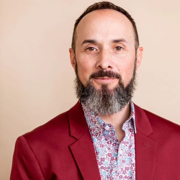 Paul Treanor (he/him/his) in a red suit, floral shirt, with long beard, brown eyes, and short hair