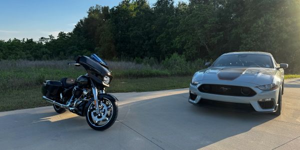 A black motorcycle and a silver sports car parked on a driveway near greenery.