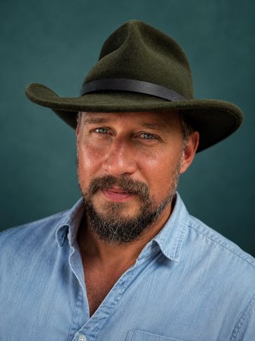 Man with a beard wearing a green hat and denim shirt.