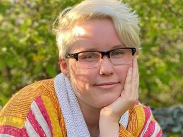 Person with short blonde hair and glasses, resting their face on their hand.