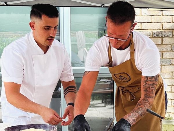 Two men cooking together outdoors, one in white chef's coat, the other in apron with tattoos.