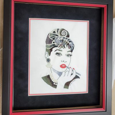 Framed embroidery of a woman applying lipstick.