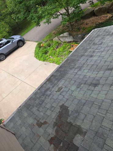 View of a driveway, parked car, green garden, and a shingled roof with shadows.