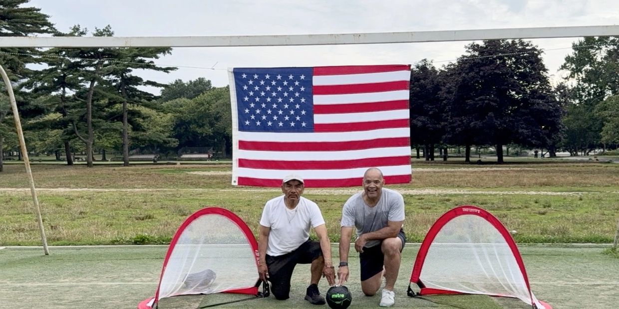 Two men pose with a soccer ball and small goals in front of an American flag.