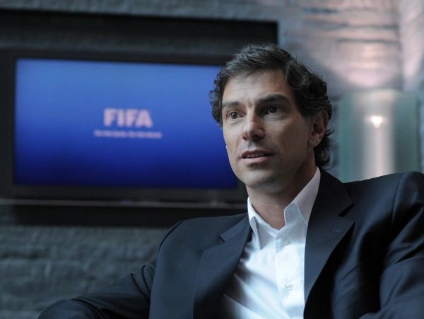 Man in a suit sitting with FIFA logo on screen behind him.