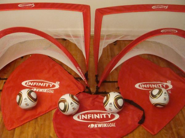 Two red pop-up soccer goals with balls and carrying bags on wooden floor.