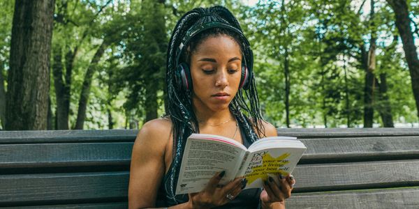 A Black Women wearing headphones sitting on a park bench reading.