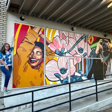 Woman standing next to a vibrant multi-panel mural featuring a joyful person and floral designs.