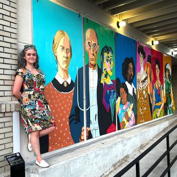 Woman stands beside colorful mural of diverse historical and cultural figures.