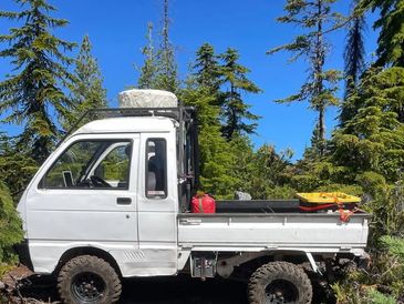 A small white off-road truck parked in a forest under clear blue sky.