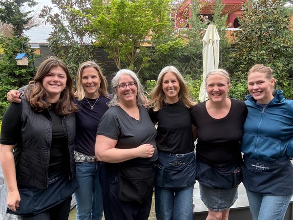 Six women smiling outdoors in casual attire with aprons.