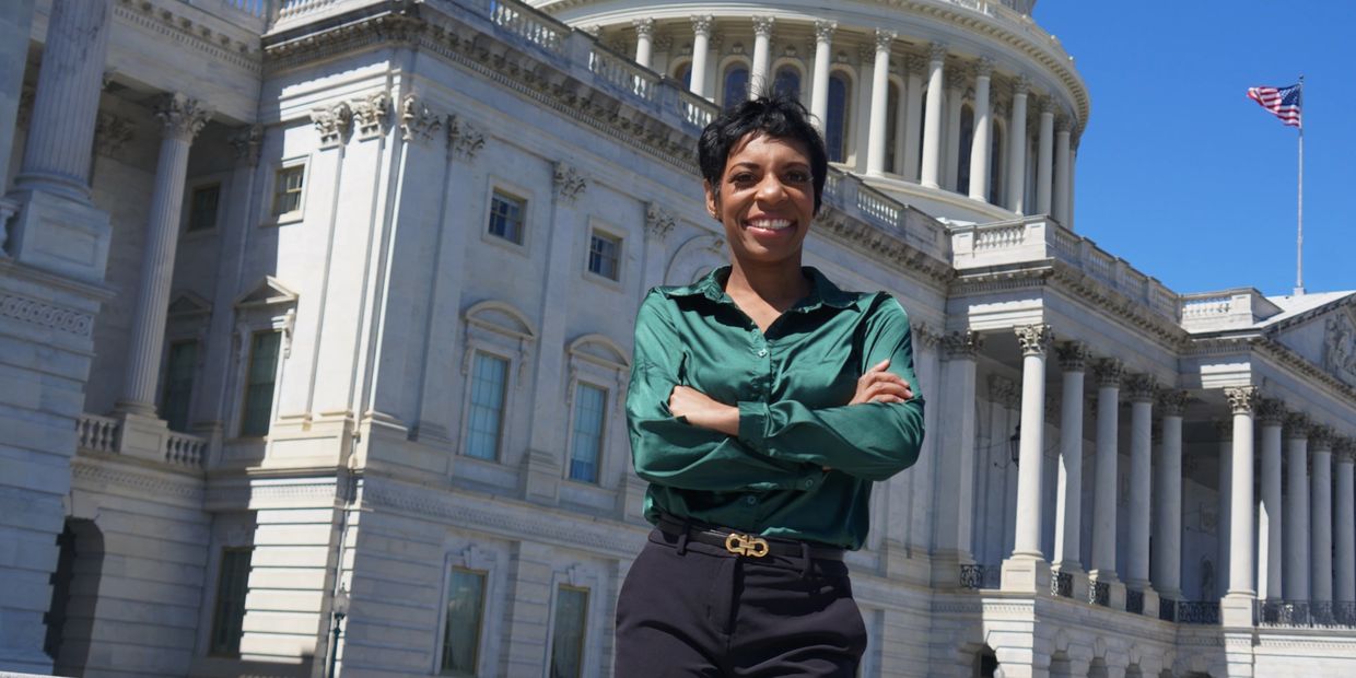 A woman stands confidently in front of the U.S. Capitol building, smiling with arms crossed, wearing