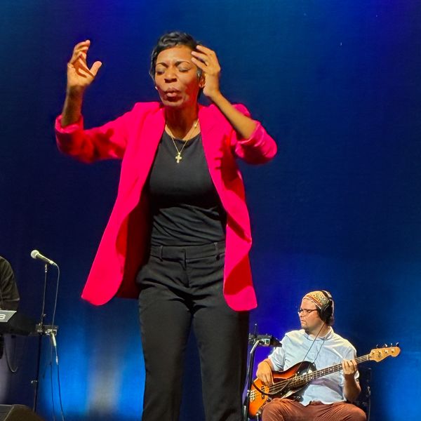 A woman in a pink blazer Interpreting on stage,  a musician with a guitar is seated behind her.