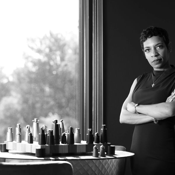 A black-and-white picture of a woman standing with arms crossed by window; a chess set on the table.