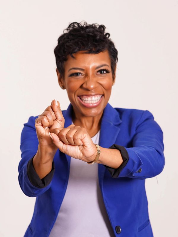 Smiling woman in blue blazer poses against plain background, hands crossed in expressive gesture.