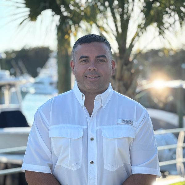Man smiling in white shirt by a marina with boats and palm trees.