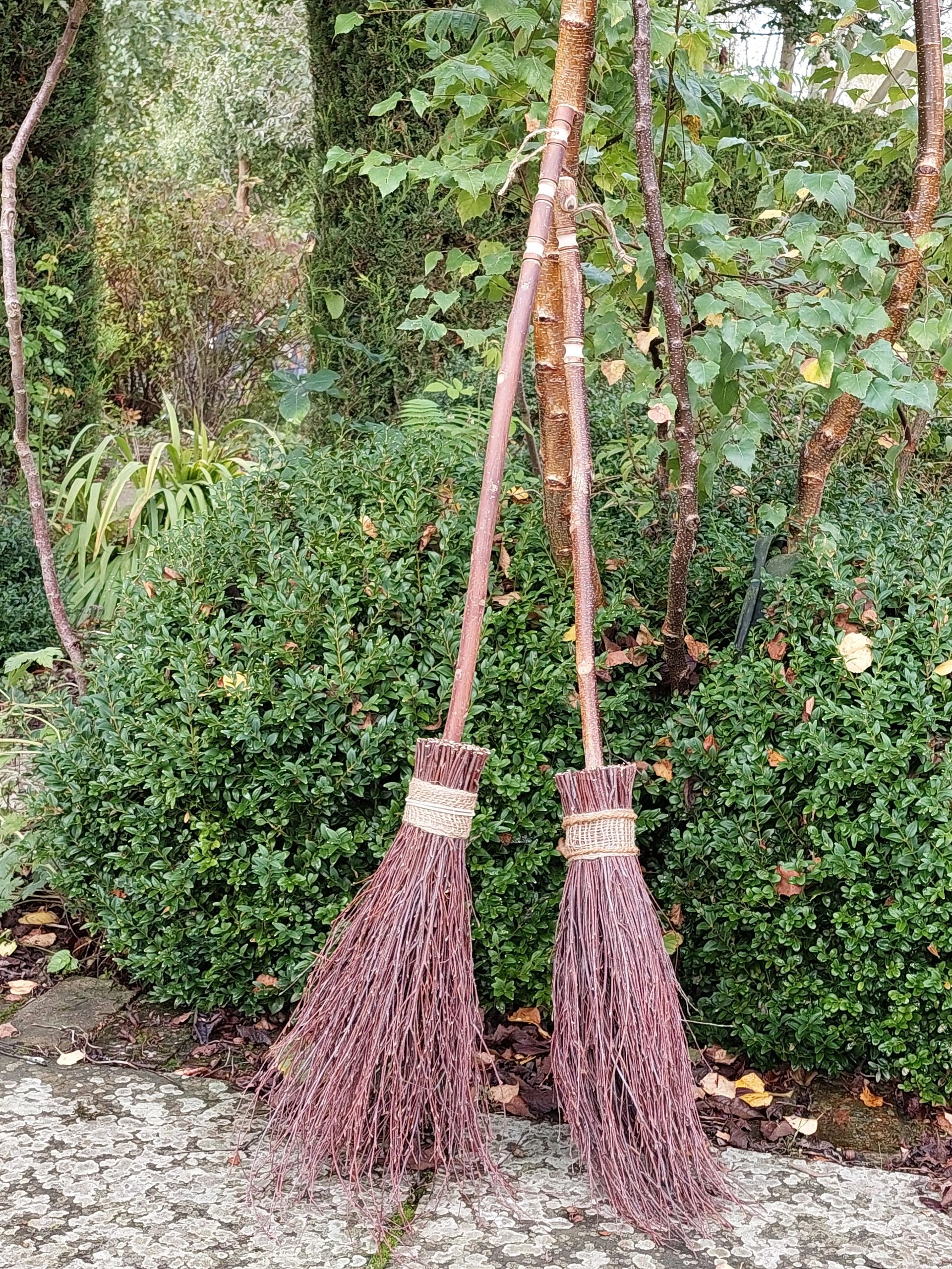 Two handmade twig brooms leaning against green bushes outdoors.