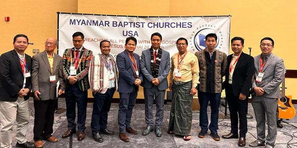 Group of men standing in front of Myanmar Baptist Churches USA banner.