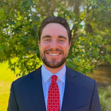 Smiling man in a suit and red tie outdoors in front of greenery.