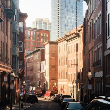 Sunlit urban street with brick buildings and parked cars.