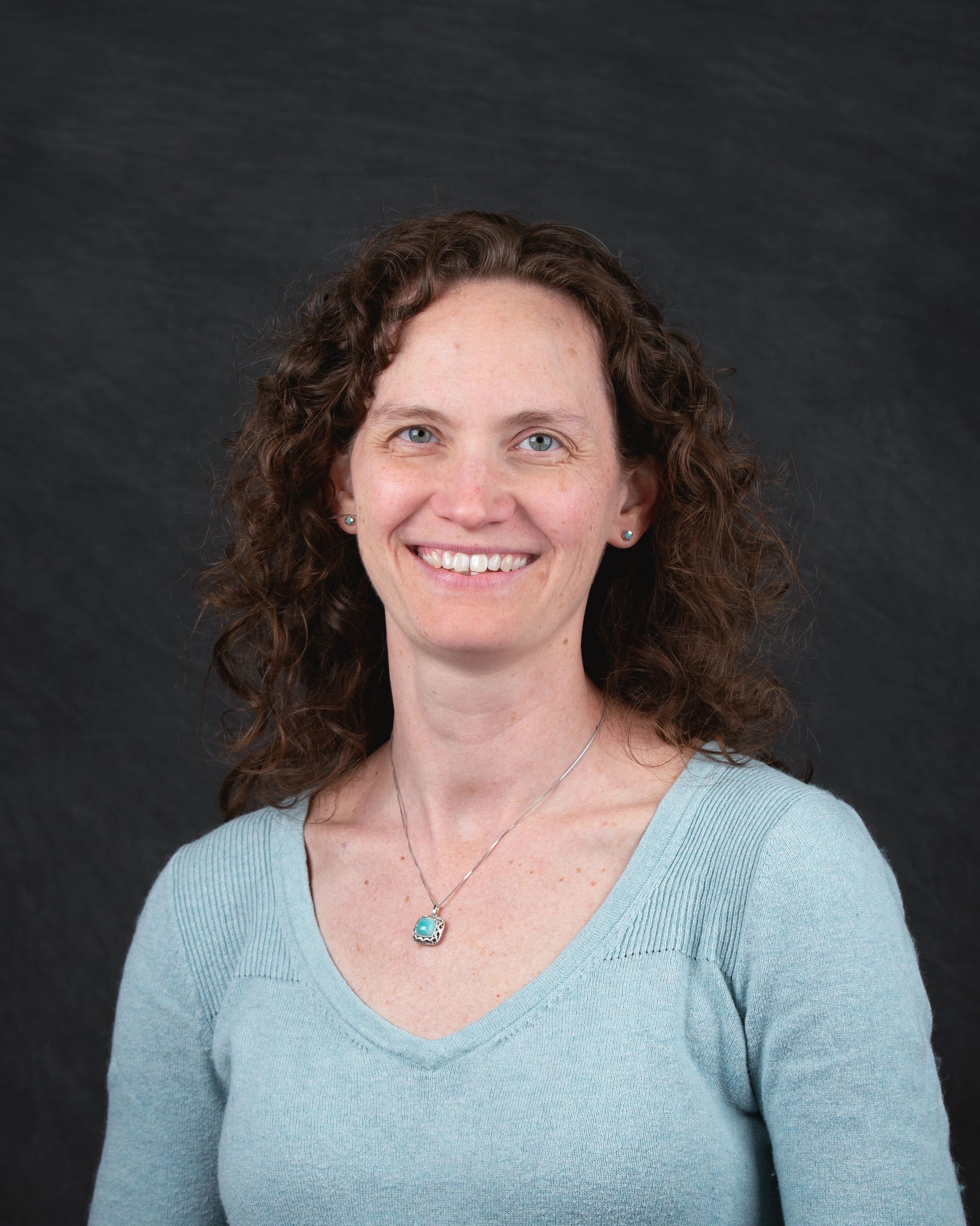 Smiling woman with curly hair wearing a light blue sweater and turquoise pendant.
