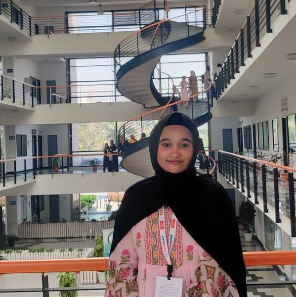A woman in a hijab stands indoors with a spiral staircase in the background.
