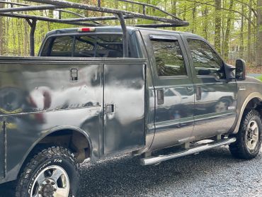 A black utility truck with a ladder rack parked on gravel in a wooded area.