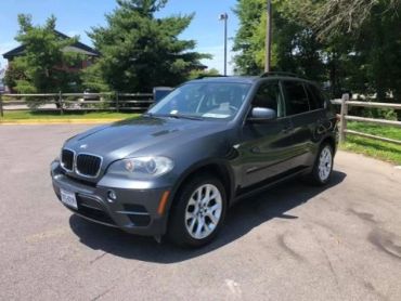 A gray BMW X5 SUV parked on a sunny day near trees and a wooden fence.