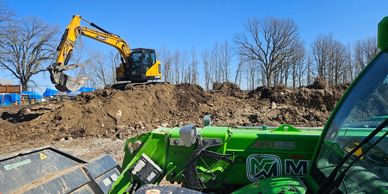 Yellow excavator on dirt mound with green machinery in foreground under clear blue sky.