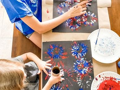Two kids creating colorful handprint art on black paper using red, white, and blue paint.