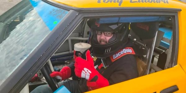 Race car driver in helmet and gloves holding a drink inside a yellow race car.