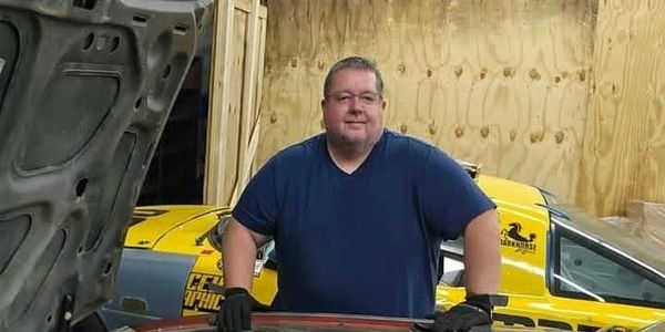 Man in blue shirt working on a car with a yellow vehicle in the background.