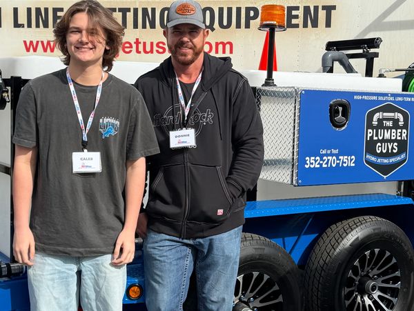Two men stand in front of a plumbing equipment trailer.