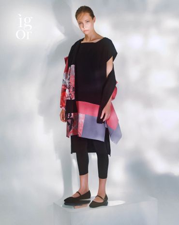 Model wearing a modern, asymmetrical dress with black, red, and purple tones in a minimalist setting.
