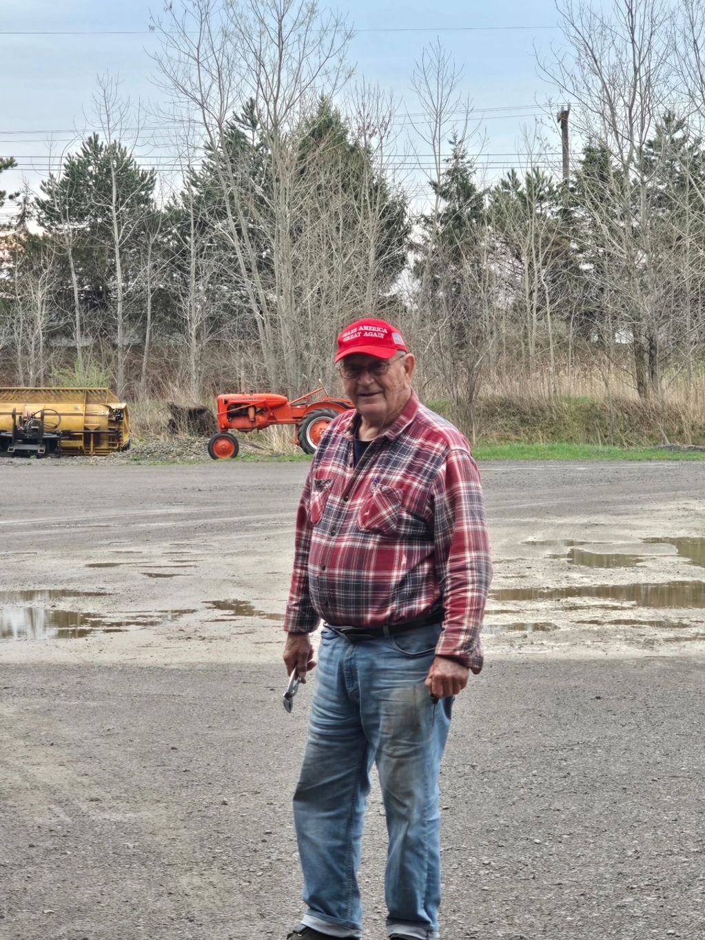 Elderly man in a red plaid shirt and cap stands outdoors near a tractor.