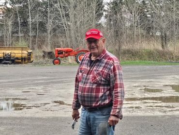 Elderly man in a red plaid shirt and cap stands outdoors near a tractor.