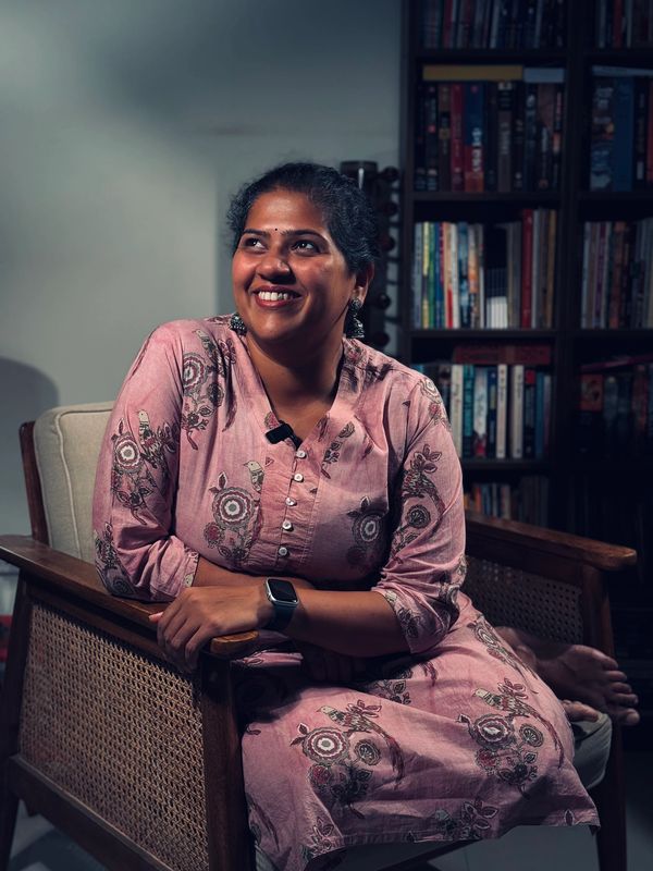 Woman smiling while seated in a wooden chair near a bookshelf.