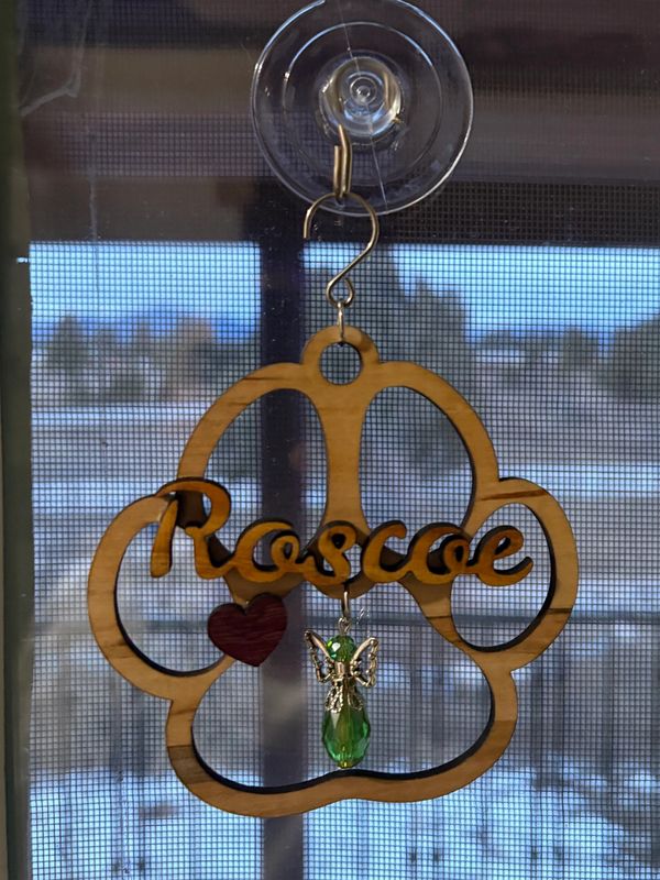 Wooden paw print ornament with the name Roscoe and a green angel charm hanging on a window.