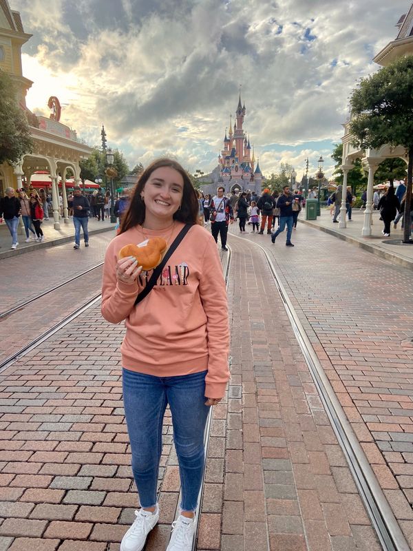 A cheerful scene at Disneyland Paris, featuring a woman enjoying a Mickey-shaped snack while strolling Main Street toward the iconic Sleeping Beauty Castle