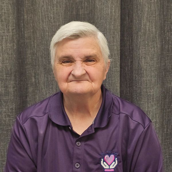 Elderly woman with white hair wears a purple shirt and smiles gently.