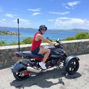 Person in red sleeveless shirt on a three-wheeled motorcycle by the seaside.