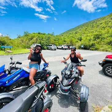 Two women enjoying a sunny day on three-wheeled motorcycles in a scenic mountainous area.