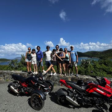 Group of nine people posing with three-wheeled motorcycles on a sunny day.