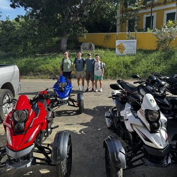Four people posing behind three colorful three-wheeled motorcycles on a sunny day.