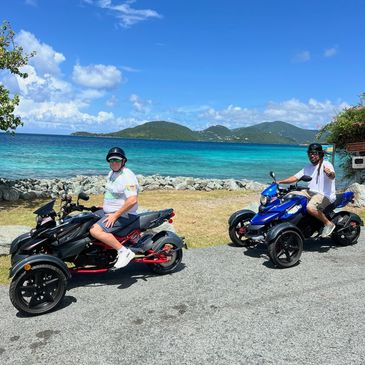 Two men on three-wheeled motorcycles near a scenic ocean view with hills in the background.