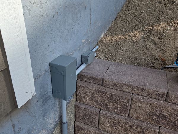 Electrical boxes mounted on a concrete wall next to a stone retaining wall.