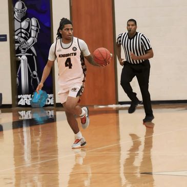 Basketball player dribbling while a referee runs behind him on a gym court.