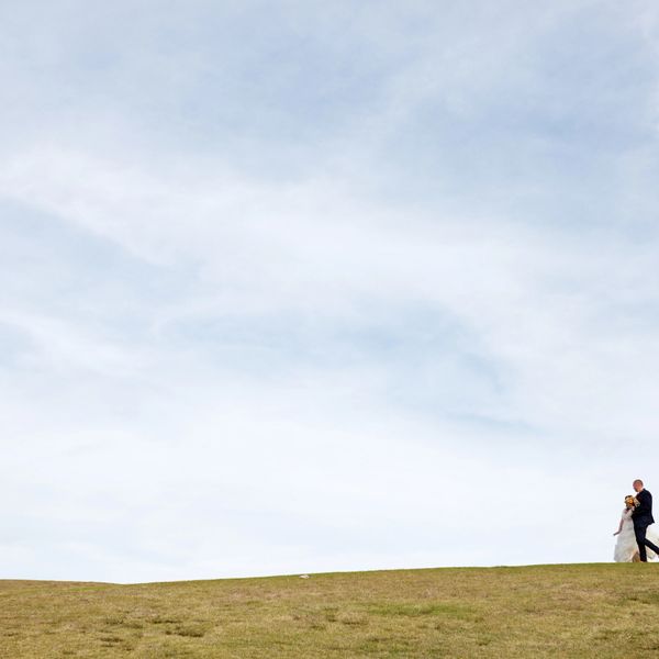 A newlywed couple walking on a grassy hill under a vast sky.